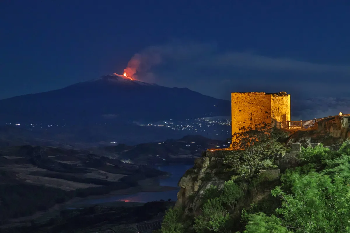 Una torre illuminata del castello di Agira di notte, con un vulcano in eruzione sullo sfondo.